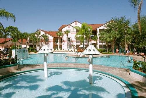 Outdoor pool area with people swimming and lounging. There are palm trees, a large circular pool, and a building with red roofs in the background.
