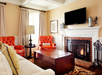 Cozy living room with a brick fireplace, two red armchairs, a wooden coffee table, and a TV mounted above the mantle. Beige walls and curtains frame the scene.