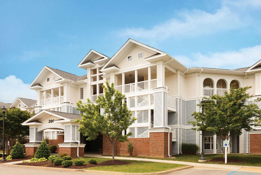 A three-story residential building with a mix of white siding and red brick, featuring balconies and gable roofs, surrounded by trees and a clear blue sky.