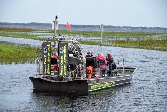 A black air boat full of people in the water with marsh grass in front of them and trees in the distance at Boggy Creek Airboat Adventures.