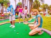 Four young adults playing mini golf outdoors, with one person preparing to putt and the others reacting and watching near a colorful structure and palm trees.