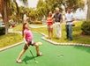 A young girl prepares to putt on a mini-golf course while three people watch her on a sunny day surrounded by palm trees.
