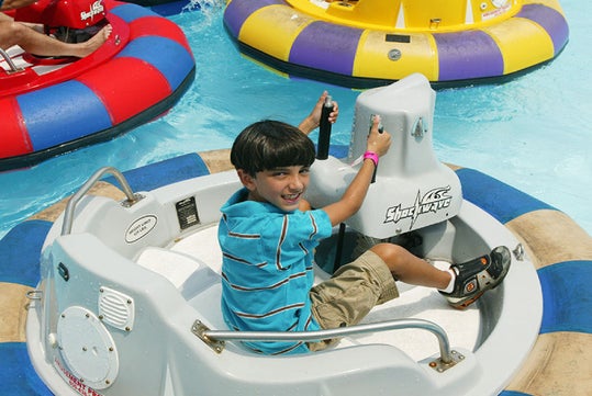 A young boy in a striped shirt rides a white bumper boat in a pool, smiling and holding the steering wheel. Other colorful bumper boats are visible in the background.