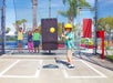 A young boy wearing a yellow helmet prepares to hit a baseball in a batting cage while four children cheer behind the fence.