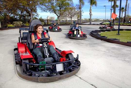 People are driving go-karts on an outdoor track with trees and buildings in the background. The person in front is smiling and turning the steering wheel.