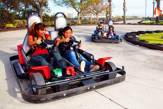 Two pairs of people drive go-karts on an outdoor track, with one kart in the foreground and another in the background. Trees and buildings are visible in the distance.