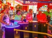 Four people play a light-up arcade game while a woman and a man watch in a brightly lit indoor arcade.
