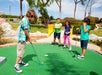 Four children are playing mini golf on a green course, with one child preparing to hit a ball while the others watch.