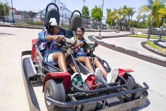 Two people drive a red go-kart together on an outdoor track, both smiling and holding the steering wheel, with palm trees and fencing in the background.