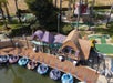 A row of colorful bumper boats docked by a whimsical, cartoon-like building near a mini-golf course in an outdoor amusement park.