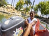 A girl smiles while steering a bumper boat on a small lake surrounded by rocks, palm trees, and greenery under a sunny sky.