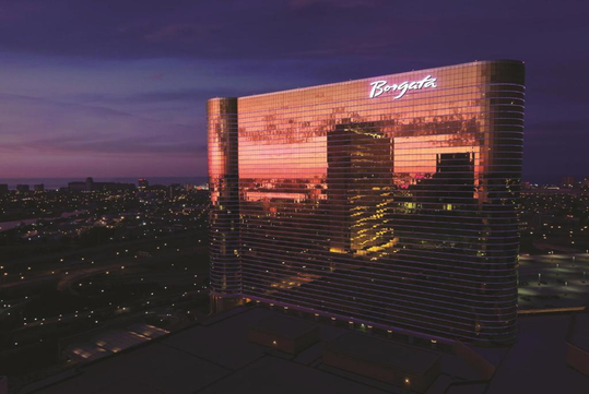 A tall, reflective building with "Borgata" on top, illuminated by a sunset, stands against a backdrop of a cityscape at dusk.