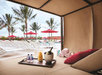Poolside cabana with a tray holding champagne, orange juice, water bottles, and glasses. Lounge chairs with red umbrellas and palm trees are visible in the background.