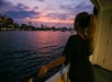 A woman with a long high pony tail leaning against the ship railing holding a glass of wine while enjoying the spectacular views of Boston at sunset.