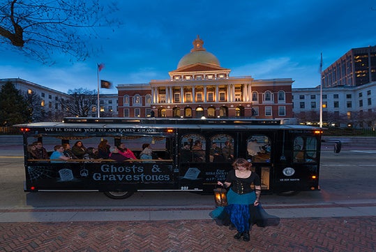 A woman in costume holding a lantern stands in front of a “Ghosts & Gravestones” tour trolley with passengers, parked by a lit government building at dusk.
