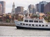 A white cruise ship with people standing on the upper and lower desk on the Boston Historic Harbor Cruise with the city in the background.