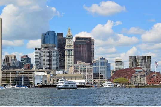 Wide shot of the Boston Harbor with a large white cruise boat in the water with the city behind it and a blue sky full of clouds overhead.