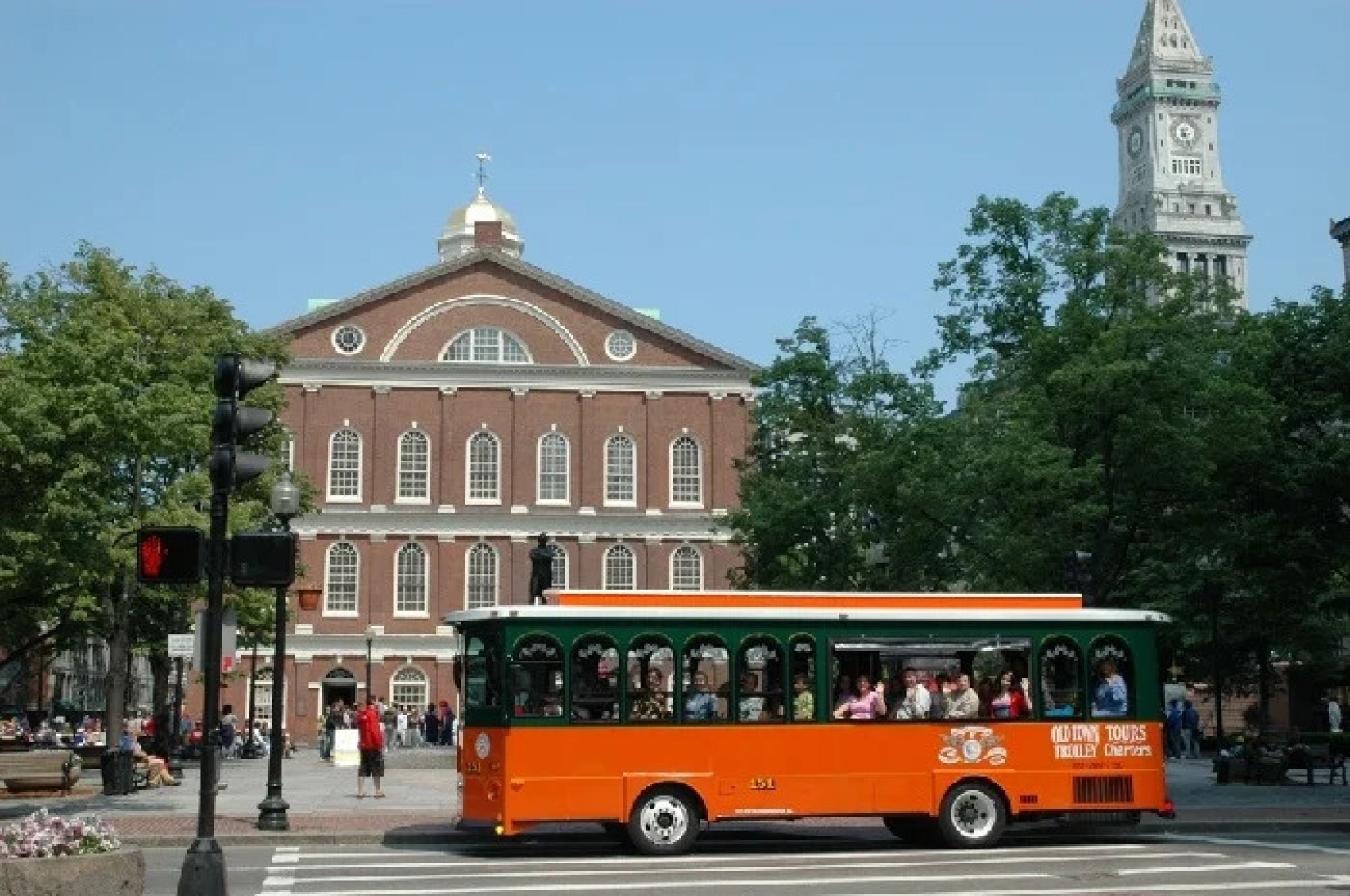 An orange and green trolley bus passes in front of Faneuil Hall in Boston on a sunny day, with people visible inside and pedestrians in the background.
