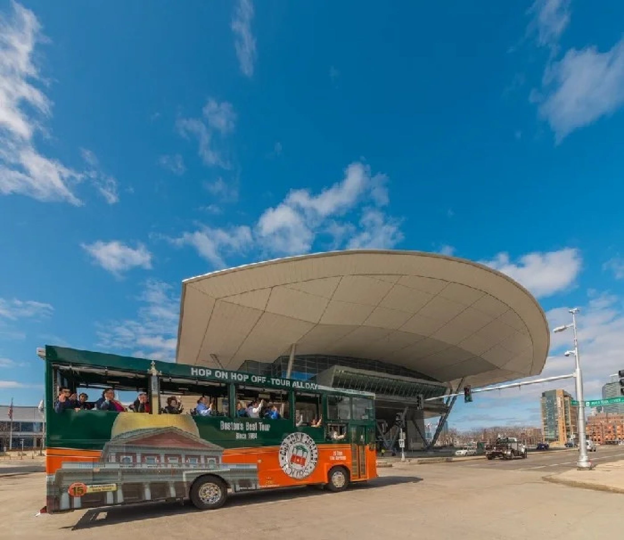A green and orange open-air tour bus drives past a modern building with a curved roof under a clear blue sky.