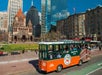 A green and orange Boston tour trolley drives past historic and modern buildings on a sunny day, with Trinity Church and skyscrapers in the background.