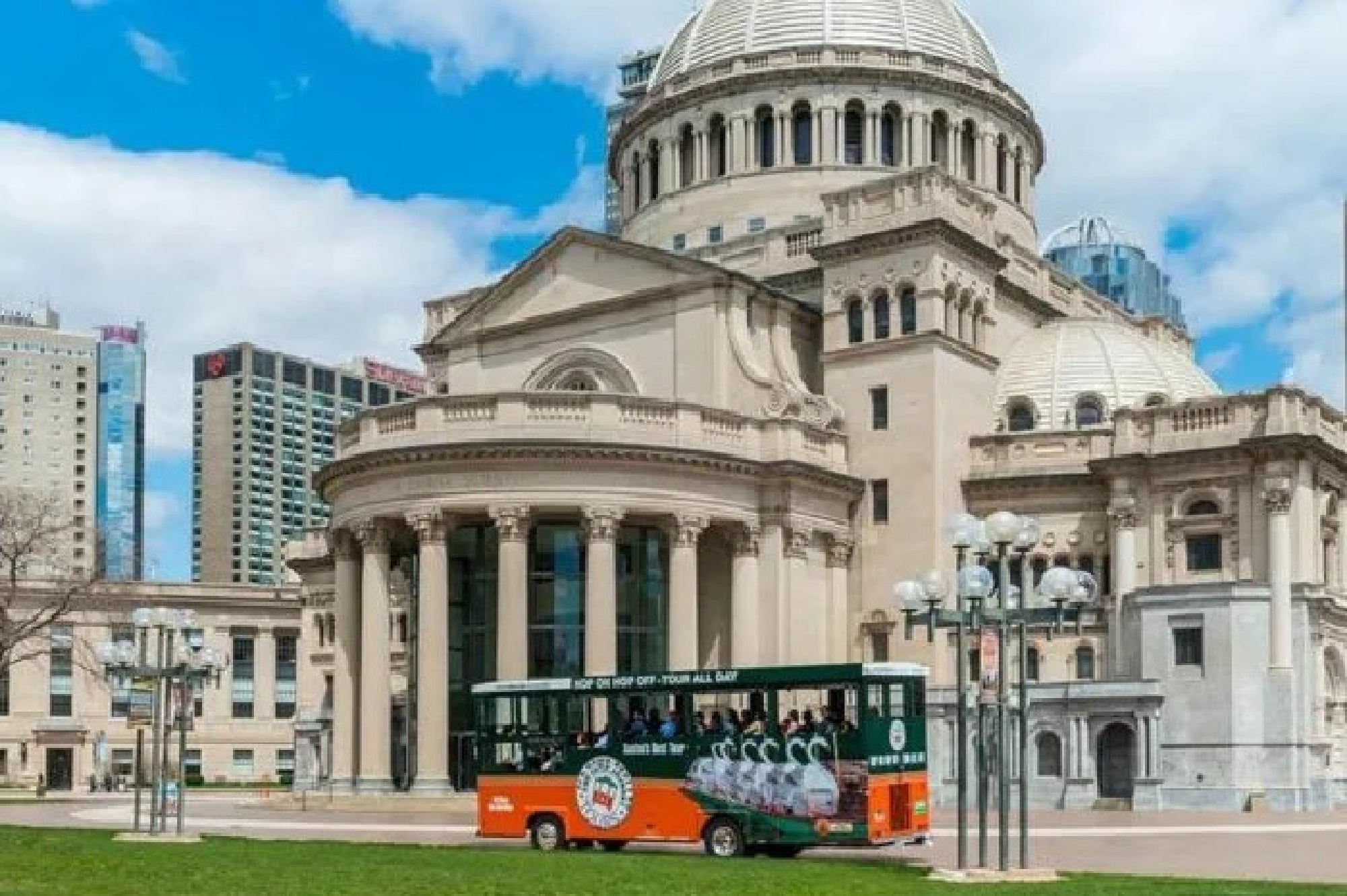 A green and orange city tour trolley passes in front of a large domed historic building with columns and ornate architecture on a sunny day.