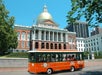An orange city tour trolley passes in front of the Massachusetts State House with its gold dome on a clear, sunny day.