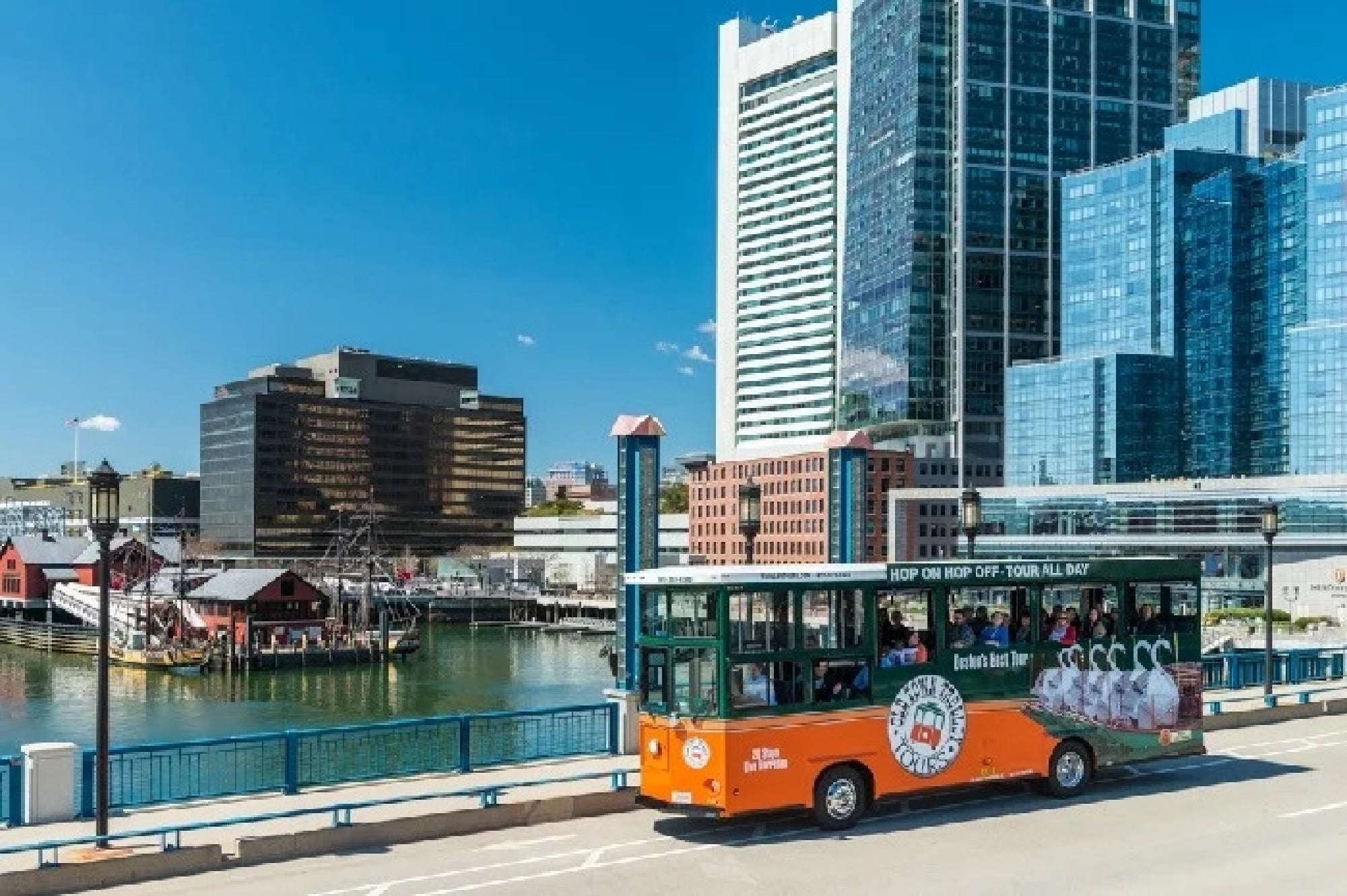 A green and orange hop-on hop-off tour trolley crosses a bridge in a modern city with water, historic ships, and tall glass buildings in the background under a clear blue sky.