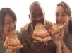 Three people are smiling and eating large slices of cheese pizza while looking at the camera.