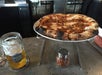 A glass of light beer, a pizza on a raised stand, and a shaker of crushed red pepper sit on a concrete bar counter.