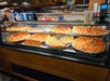 A display case with various whole and sliced pizzas, including pepperoni and other toppings, in a restaurant setting with staff visible in the background.