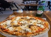 A baked pizza with cheese and sausage sits on a wooden table next to a bottle of Peroni beer, red pepper flakes, and grated cheese shakers in a restaurant setting.
