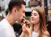 A woman holds a chocolate-covered pastry while a man takes a bite; both appear to be outdoors and are smiling.