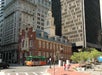 Historic brick building with a clock tower stands among modern skyscrapers; a green and orange trolley and pedestrians are visible on the street in front.