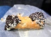 A partially eaten cannoli with chocolate chips on both ends sits on wax paper, with cream filling visible inside the pastry shell.