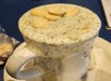 A white mug overflowing with creamy soup, possibly clam chowder, topped with several oyster crackers and herbs, sits on a saucer with soup spilling over the sides.