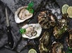 Opened and unopened oysters on a dark surface with ice, seaweed, lime slices, parsley garnish, and an oyster knife nearby.