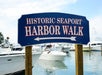 A blue sign reads "Historic Seaport Harbor Walk" with boats docked in water and a partly cloudy sky in the background.