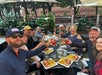 A group of people sit at an outdoor restaurant table, raising their drinks and smiling, with plates of food and greenery in the background.