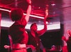 Three people are dancing indoors under red lighting, with one woman in the center smiling and reaching her hand up.