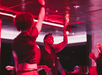 People are dancing in a dimly lit room with red lighting. One person raises a hand in the foreground.