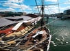 A tall ship is docked next to a red building on a river, with people on board and floating crates in the water nearby under a blue sky.
