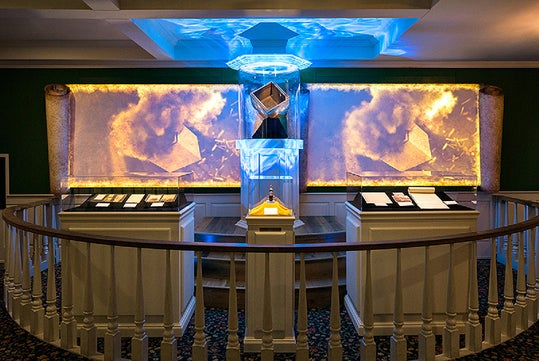 Museum exhibit displays illuminated artifacts, documents, and a suspended cube with blue lighting, surrounded by a white railing and dramatic backdrop.