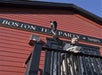 A man in historical costume stands on a balcony in front of a red building with a sign reading "Boston Tea Party Ships," raising his hat.