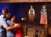A man and two children observe two animated women dressed in historical clothing displayed on a screen or hologram in a themed exhibit.