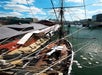 A tall ship docked at a pier on a sunny day, with people on deck and floating crates or barrels in the water nearby.