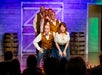 Actors in period costumes perform a musical number on stage in front of an audience, with a wooden barn backdrop and barrels on either side.