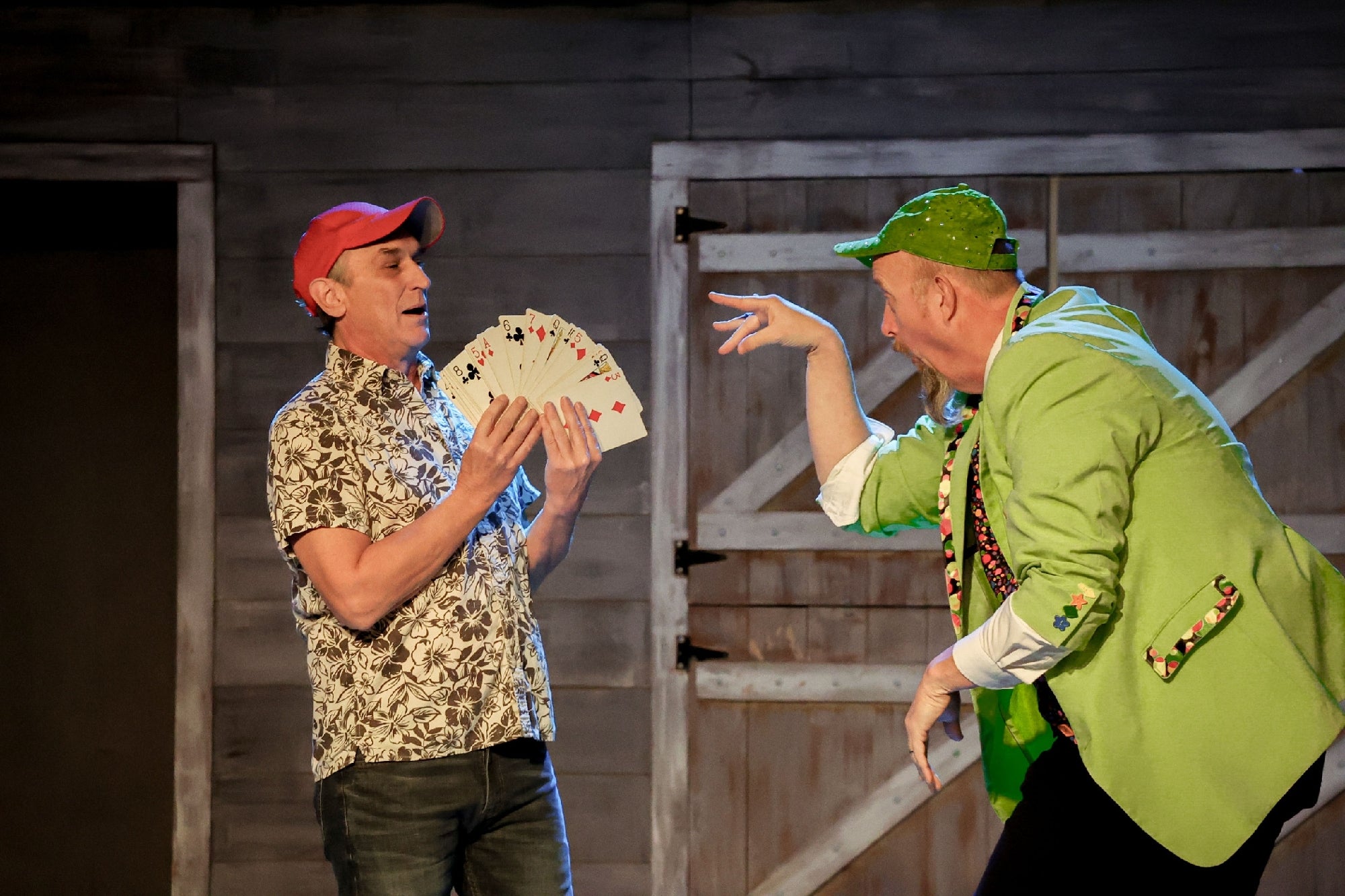Two performers on stage: one in a floral shirt and red cap holds playing cards, while the other in a green jacket and cap gestures animatedly. A wooden backdrop is visible.