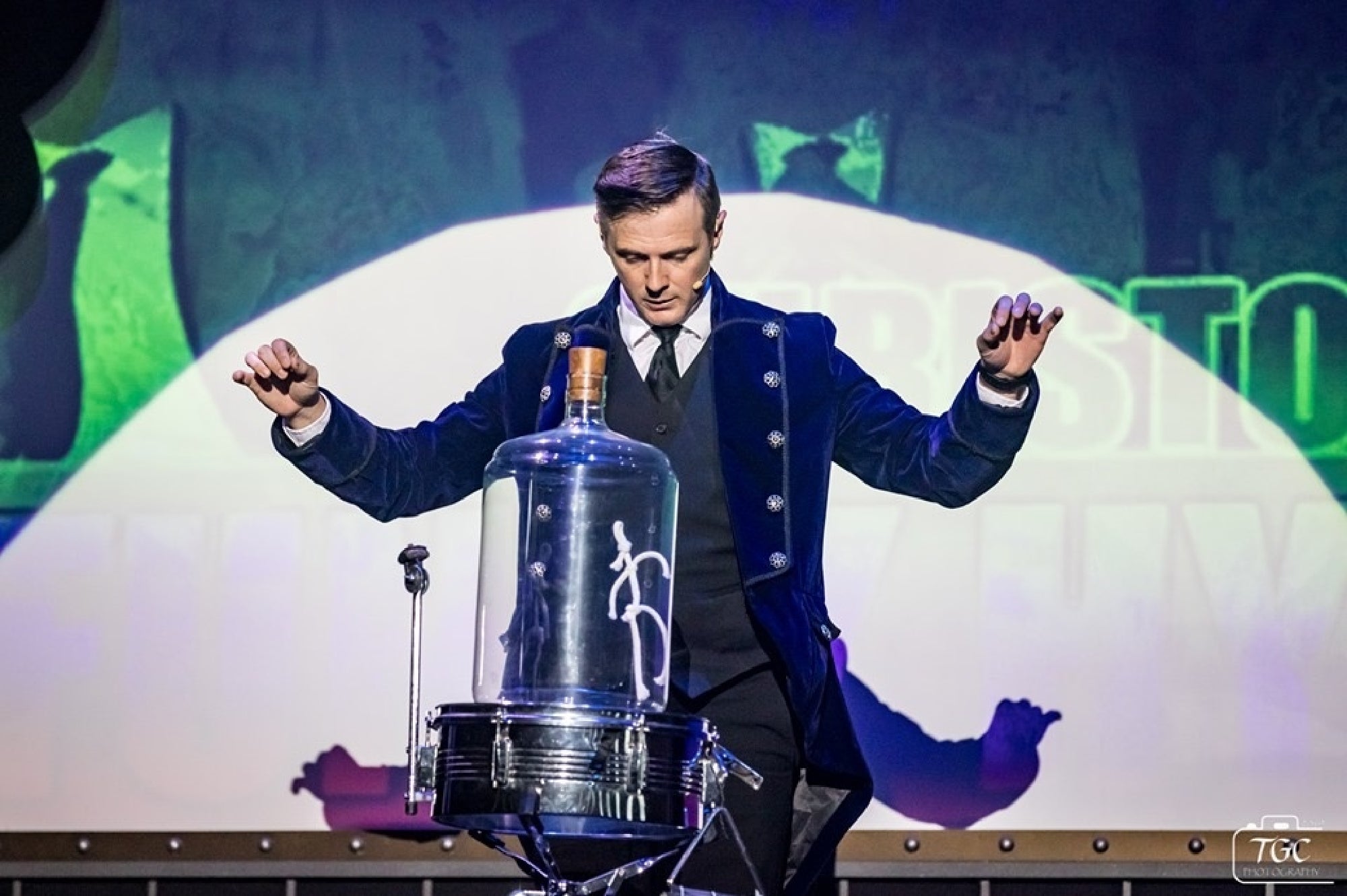 A magician in a dark coat stands with raised hands behind a glass dome on a drum, performing on a stage with dramatic lighting.