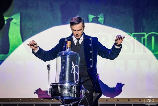 A magician in a dark coat stands with raised hands behind a glass dome on a drum, performing on a stage with dramatic lighting.