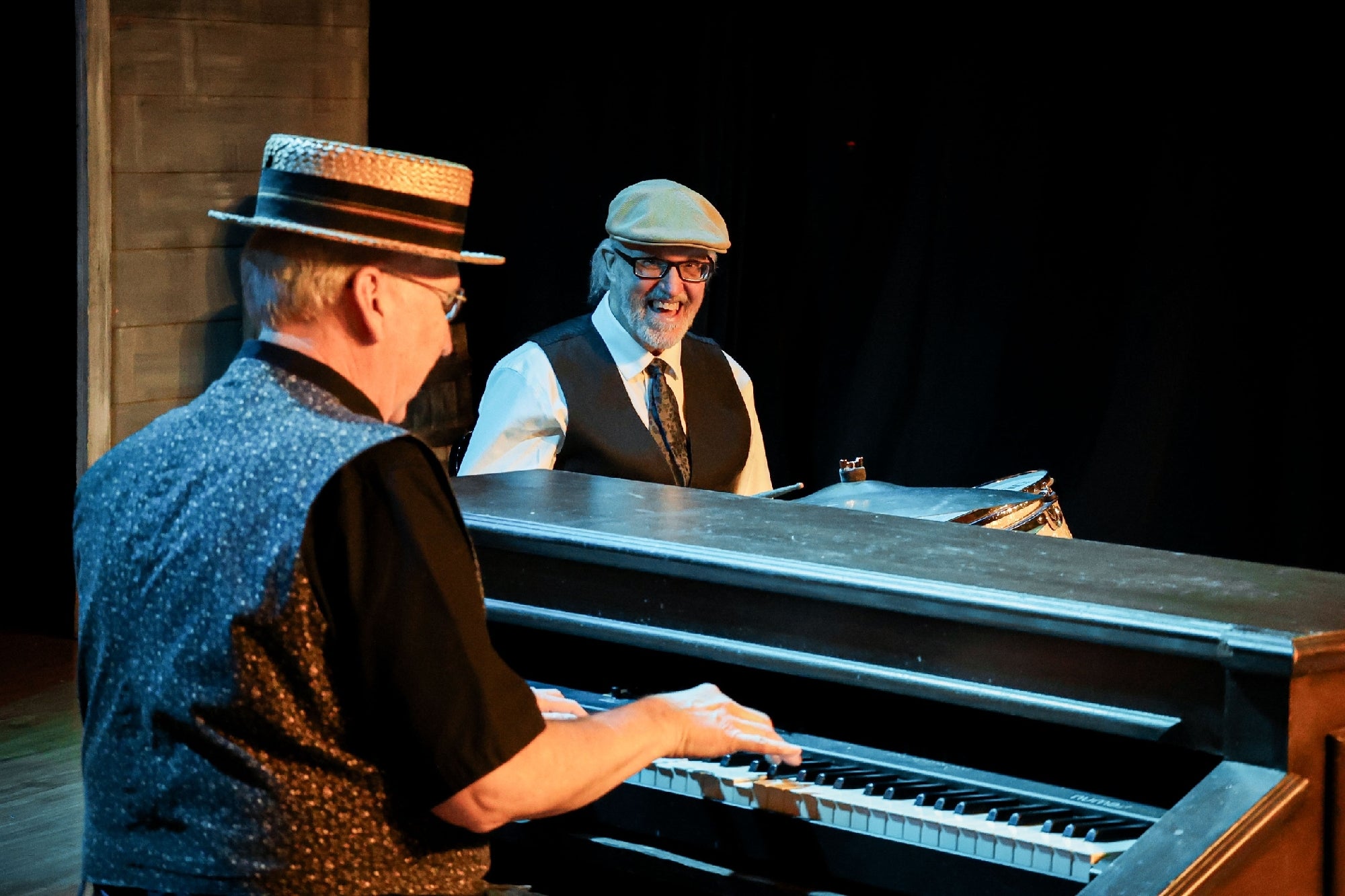 Two men wearing hats and vests play piano and drums on a dimly lit stage.